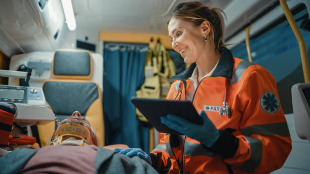 Female EMS Professional Paramedic Using Tablet Computer to Fill a Questionnaire for the Injured Patient on the Way to Hospital. Emergency Care Assistant Comforting the Patient in an Ambulance. Female EMS Professional Paramedic Using Tablet Computer to Fill a Questionnaire for the Injured Patient on the Way to Hospital. Emergency Care Assistant Comforting the Patient in an Ambulance. paramedic photos stock pictures, royalty-free photos & images