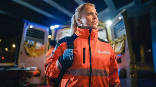 Portrait of a Female EMS Paramedic Proudly Standing in Front of Camera in High Visibility Medical Orange Uniform with "Paramedic" Text Logo. Successful Emergency Medical Technician or Doctor at Work.