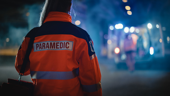 Paramédica de EMS con orgullo de pie con su espalda se volvió a la cámara en uniforme de alta visibilidad médico naranja con logotipo de texto 