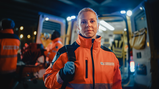 Retrato de una mujer paramédica de EMS orgullosamente de pie frente a la cámara en uniforme de alta visibilidad médica naranja con logotipo de texto 
