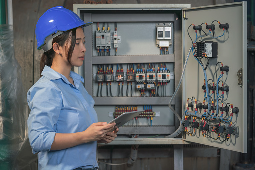 asian female industrial electrical engineer with safety hardhat and tablet on hand working in factory checking and maintenance electric control switchboard