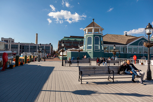 Alexandria, VA, USA 11-28-2020: A sunny day on the waterfront of the old town district of Alexandria with people sitting on benches on the pier in front of the lighthouse. There are docked vessels.