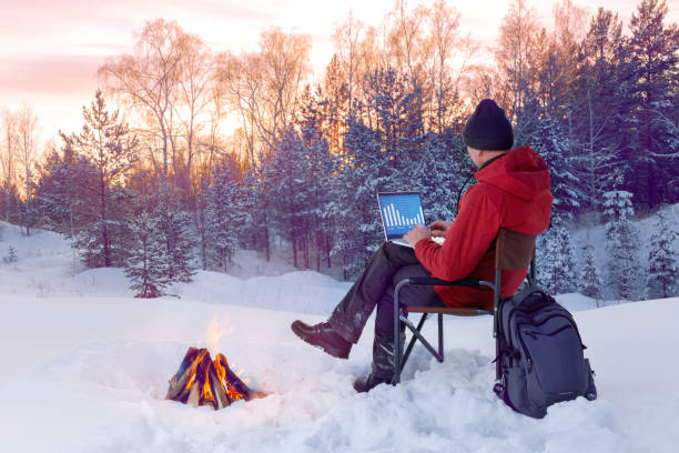 istock Man using laptop on deck chair near the fire at winter forest. Remote work. 1291121160