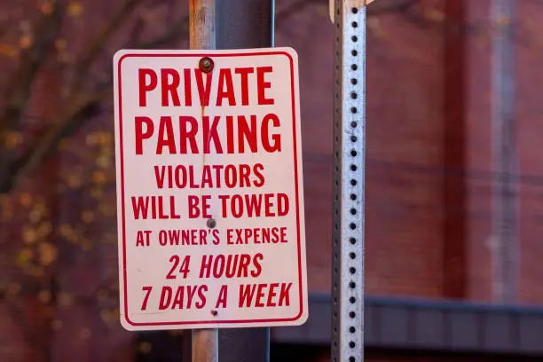 A metal sign at a parking lot that says " Private Parking, violators will be towed at owner's expense 24 hours seven days a week. " A white metal plate with red text attached on a post with screws.