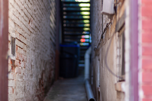An abstract out of focus image of a narrow alley between brick walls of two vintage buildings at an urban location. The paint on walls is chipped, there is a water pipe, staircase, trash can and wires