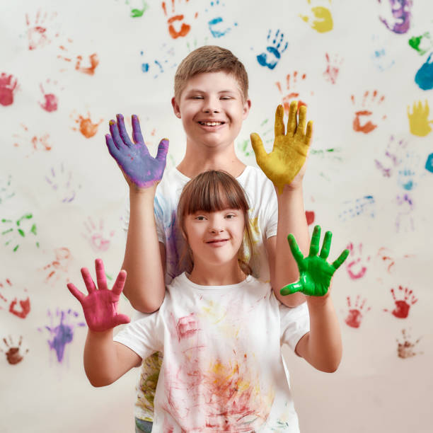 gosses heureux, garçon et fille handicapés avec le syndrome de down souriant à l’appareil-photo, affichant leurs mains peintes dans les peintures colorées pour des impressions de main sur le mur - child cheerful little girls down syndrome photos et images de collection