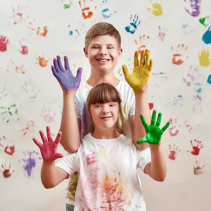 Happy kids, disabled boy and girl with Down syndrome smiling at camera, showing their hands painted in colorful paints for hand prints on the wall. Children with disabilities and special need concept