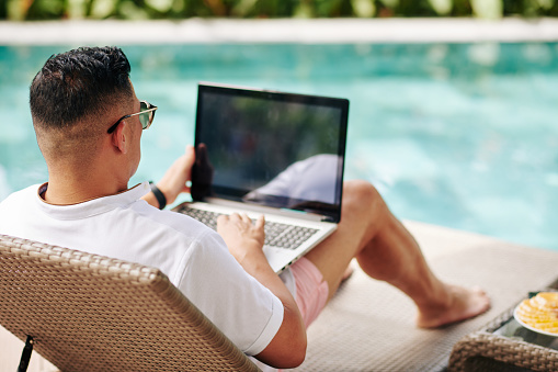Man in glasses lying in chaise-lounge by swimming pool and checking e-mails on laptop