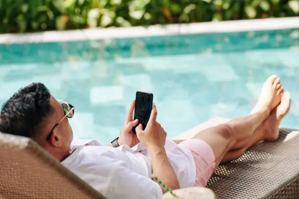 Man spending sunny day by swimming pool and using mobile application or checking social media on smartphone