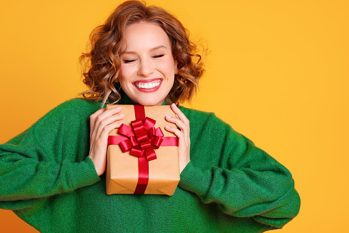 cheerful funny surprised young woman laughs in a green sweater with closed eyes holds a Christmas gifts on a colored yellow background