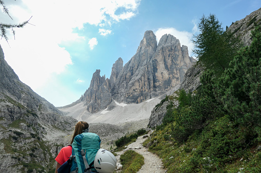A woman with big backpack and sticks, hiking in high Italian Dolomites. There are many sharp peaks in front of her. She is going up. There are a few trees around. Sunny day. Outdoor exercising