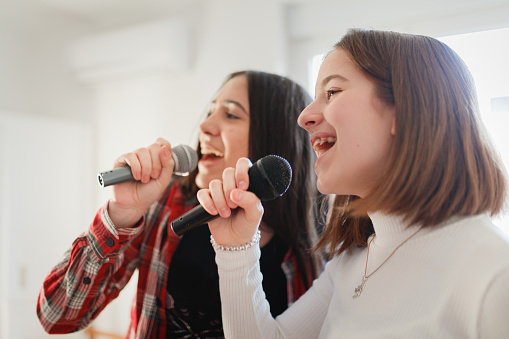 Two cheerful teenage girls singing on wireless microphones in duet, pretending to perform on concert, having fun together at home in COVID-19 pandemic lockdown