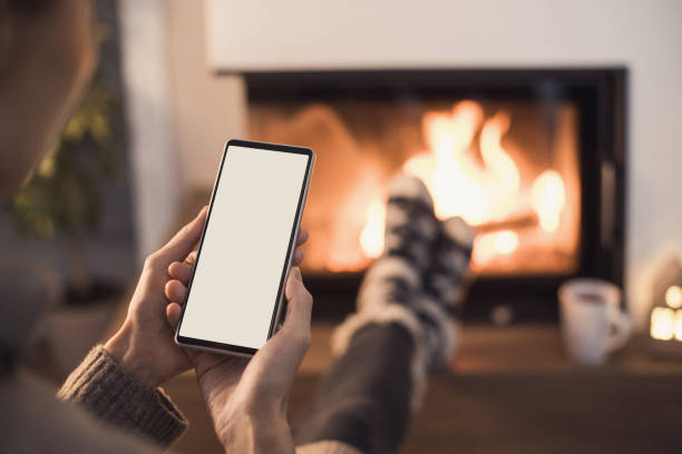 Young woman using smartphone near the fireplace at home, white blank empty screen Female holding mobile phone in winter. Connection, communication, mobile apps, technology, winter lifestyle, Christmas time, mock-up concept modern fireplace screen stock pictures, royalty-free photos & images