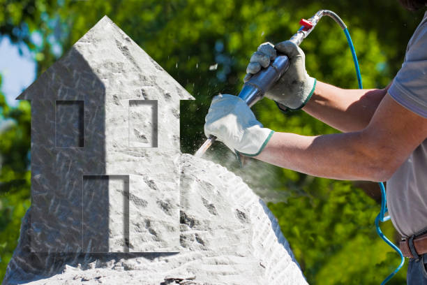 istock Man at work with compressed air chisel and protective gloves carving a little house on a stone block - concept image - It is not a real image it is a photomontage 1291163204