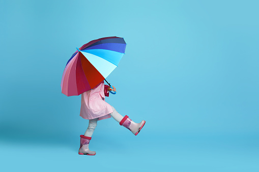 funny little child girl with multicolored umbrella in pink rain coat and rubber boots on blue background. copy space