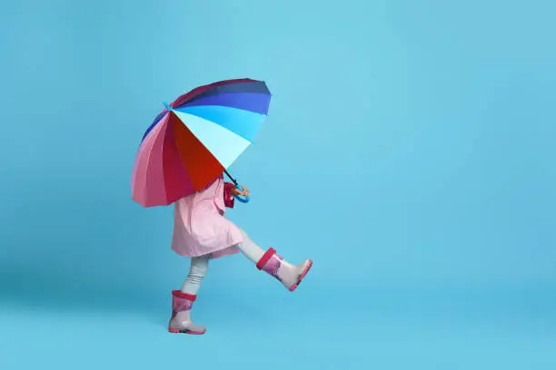 funny little child girl with multicolored umbrella in pink rain coat and rubber boots on blue background. copy space