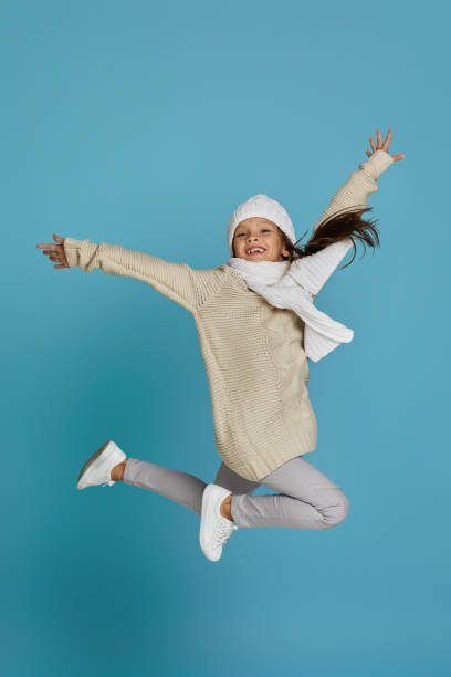 little child girl in winter knitted hat and sweater jumping Excited smiling cheerful little child girl in white winter knitted hat and sweater jumping on blue background kids winter fashion stock pictures, royalty-free photos & images