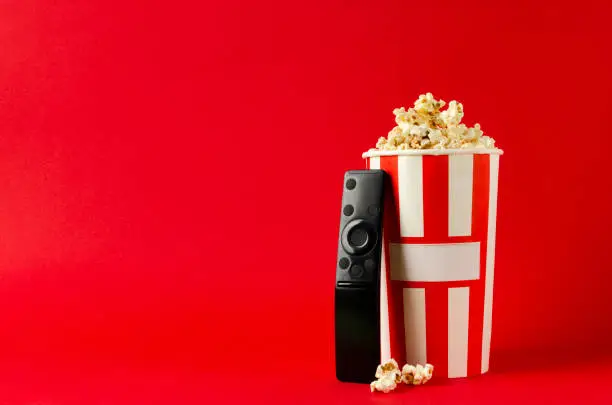 Photo of Paper box full of sweet pop corn and tv clicker on the bright red background.Empty space