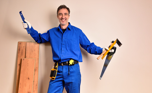 Parquet assembler in overalls smiling with tools in hands leaning on parquet slats in a room. Horizontal composition.