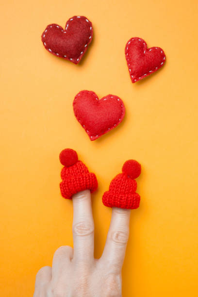 Happy valentine's day creative concept on orange. Women's fingers depict the image of two people in love in winter knitted miniature hats. Red felt heart with white stitches felt heart shape small red stock pictures, royalty-free photos & images