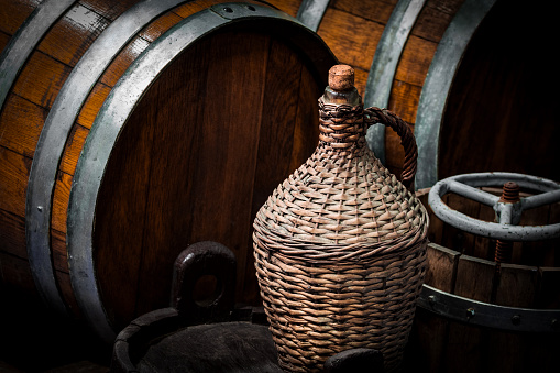 Close-up of old wicker carboy on barrels in wine cellar.