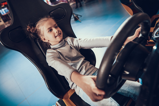 Cute Little Girl Playing Racing Simulator Game. Adorable Child at Steering Wheel of Car Simulator. Modern Technologies. Game Machine. Entertainment Concept. Cheerful Kid Enjoying Driving.