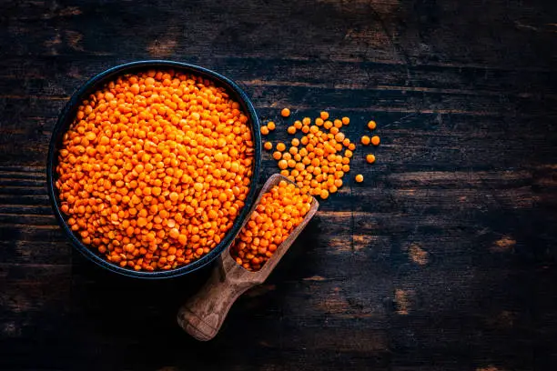 Legumes: Red lentils in a black bowl shot from above on dark wooden table. A serving scoop with lentils is beside the bowl. The composition is at the left of an horizontal frame leaving useful copy space for text and/or logo at the right. High resolution 42Mp studio digital capture taken with Sony A7rII and Sony FE 90mm f2.8 macro G OSS lens