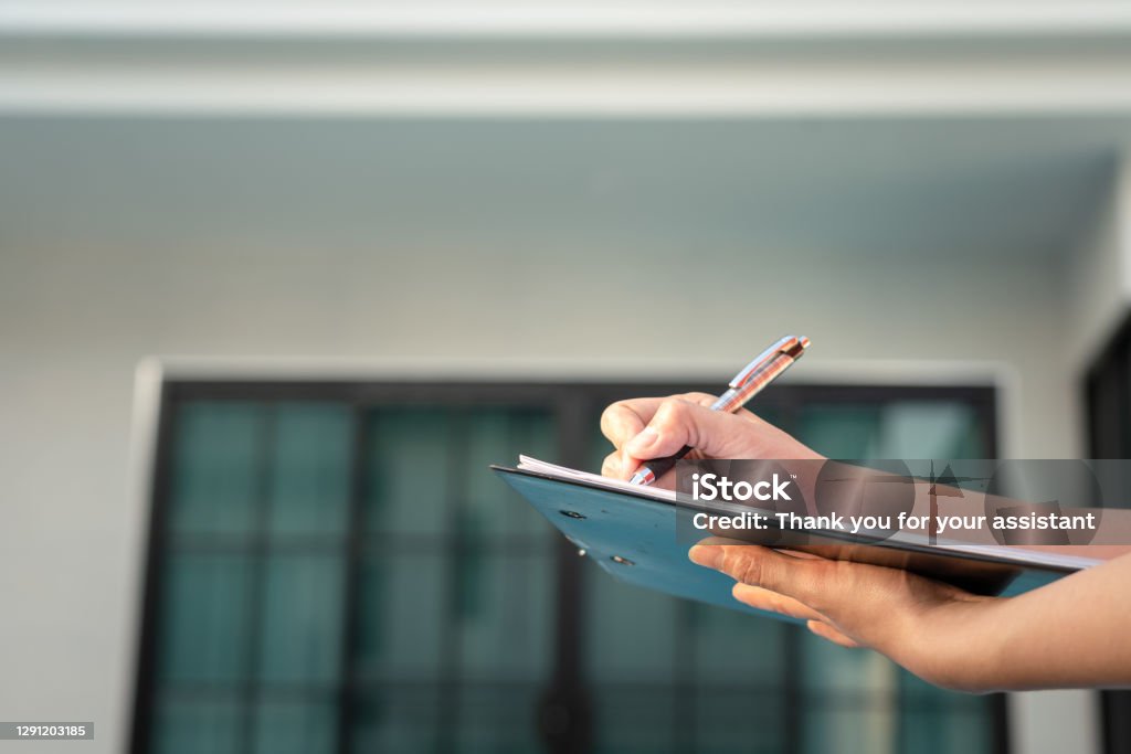 Writing down on paper for quality verify concept. Action of an engineer is writing down on the clipboard paper to verify the quality of new house that already finish (as blurred background). Home developer business and industrial photo. Quality Control Stock Photo