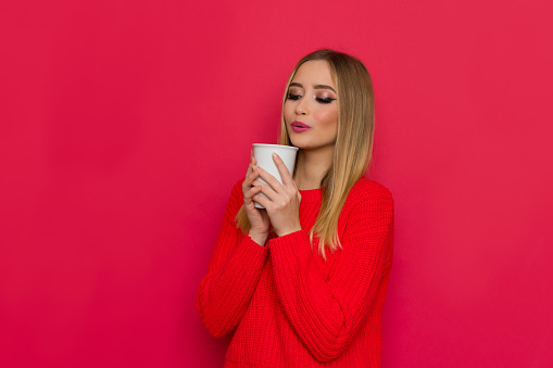 Cute woman in red sweater is holding white cup and blowing. Waist up studio shot on red background.
