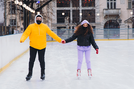 Young man in yellow jacket and a woman with curly hair wearing protective face masks feeling happiness having a bright weekend ice-skating on the ice rink together