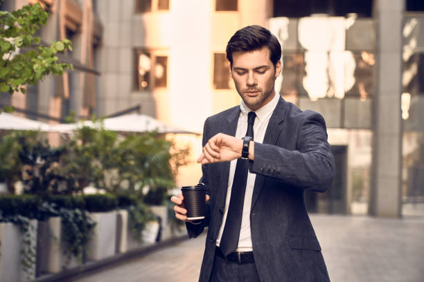 istock Confident young man in full suit holding newspaper and looking at his watch while standing outdoors with cityscape in the background 1291229356