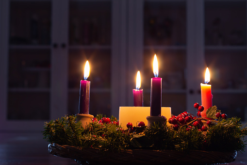 Christmas advent wreath in dark room in front of white cabinets with all four candles lit, red berries and evergreen boughs on wreath