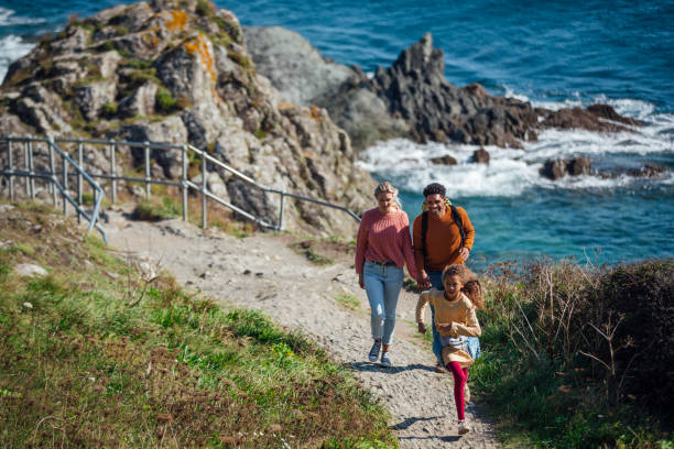 The Most Tranquil Walk A couple walking up a rock footpath along the cliff edge of Polperro, Cornwall, beside the sea. Their young daughter is running ahead in front of them. staycation stock pictures, royalty-free photos & images