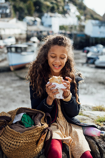 A young girl sitting on a stone wall in the fishing village of Polperro, Cornwall. She is eating a Cornish pasty while smiling.