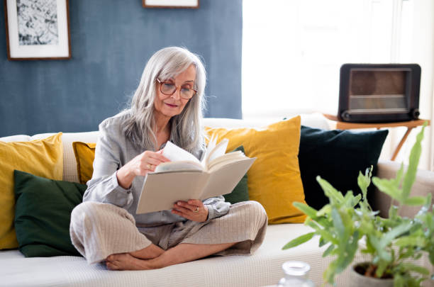 retrato de una mujer mayor sentada en el interior del sofá de su casa, leyendo libro. - women book mature adult reading fotografías e imágenes de stock