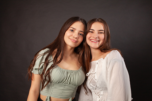 Close up studio portrait of two 17 year old teenage girls with long brown hair in summer blouses on black background