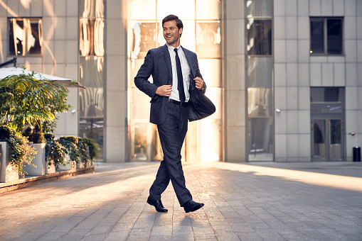 Young and successful. Handsome young businessman adjusting his jacket and looking away while walking outdoors with office building in the background.