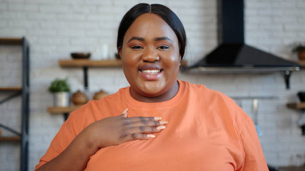 Young African-American lady in pink t-shirt speaks at camera Cheerful plump African-American lady in orange t-shirt speaks at camera moving hands actively standing against kitchen closeup huge black woman pictures stock pictures, royalty-free photos & images