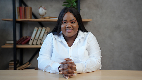 Serious body positive young African-American woman with loose dark hair in white shirt sits at table and smiles at camera closeup