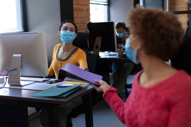 istock Diverse female colleagues wearing face masks passing file between desks 1291258052
