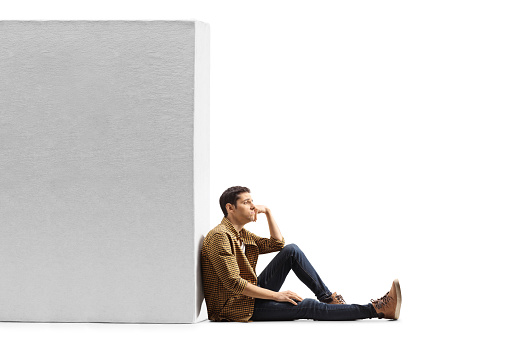 Pensive young man in casual clothes sitting on the floor and leaning on a wall isolated on white background