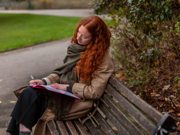 fare pubblicità a casa studiando su una panchina del parco per una giovane ragazza adulta con abbelliti capelli ricci rossi - proofreading reading and writing writing exam foto e immagini stock