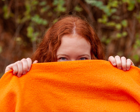 Red haired teenage girl peeking from behind an orange blanket she's holding up.
Orange blanket can be used for copy space.