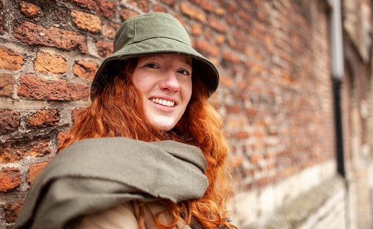 Outdoor portrait of a teenage girl with curly long red hair.\nLeans against a red brick wall on the sidewalk. Dressed in stylish coat with matching scarf.\nCopy space provided.