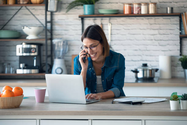 istock Beautiful young business woman talking with her mobile phone while working with computer in the kitchen at home. 1291288113