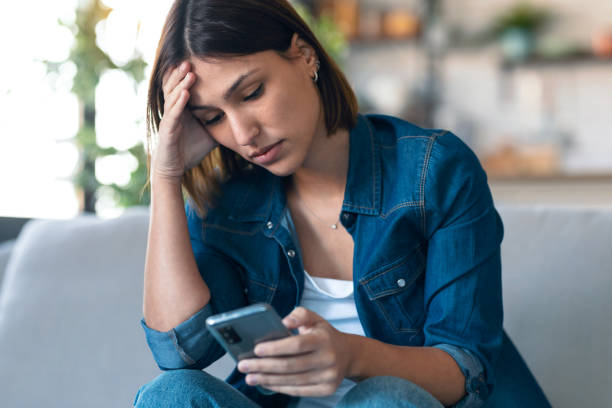 istock Worried young woman using her mobile phone while thinking about problems sitting on couch in the living room at home. 1291288735