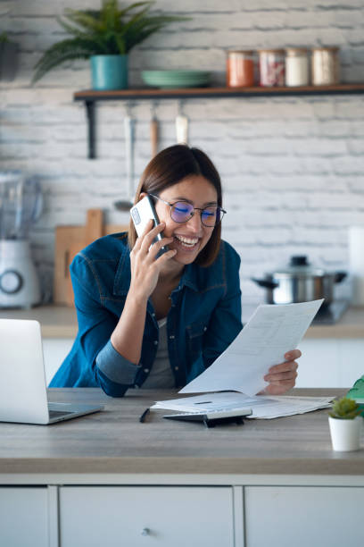 istock Beautiful young business woman talking with her mobile phone while working with computer in the kitchen at home. 1291288902