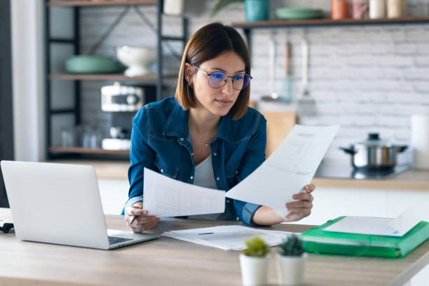 istock Pretty young business woman working with computer while consulting some invoices and documents in the kitchen at home. 1291289237