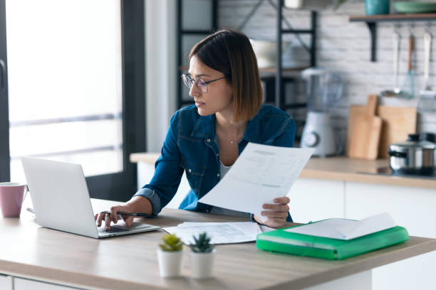istock Pretty young business woman working with computer while consulting some invoices and documents in the kitchen at home. 1291289950