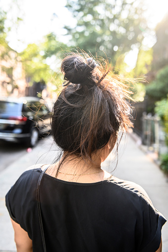 This is a photograph of the back of a Korean American woman with her hair in a topknot bun in her 30s walking on a sidewalk in Brooklyn, New York on a summer day.